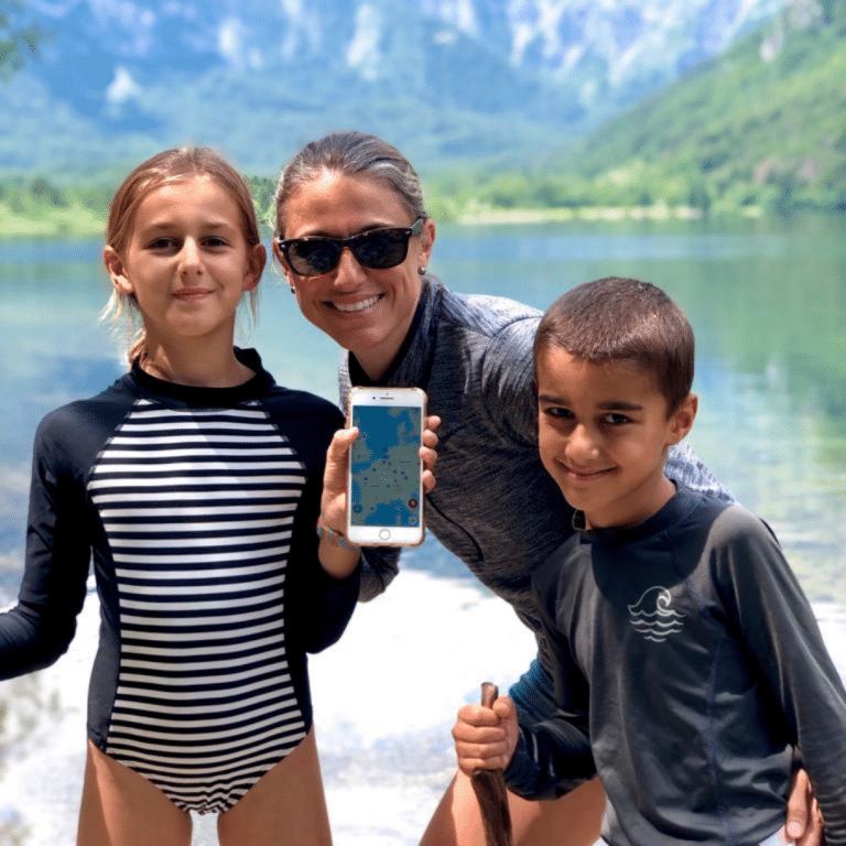 A mom with her two children in front a mountain and lake. She is holding a phone with a screenshot of the Gowhee app.