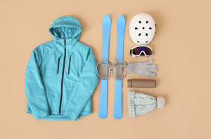 Flat lay of ski trip packing list essentials including a teal ski jacket, blue skis with bindings, white helmet, ski goggles, gloves, knit hat, and thermos on a neutral background.